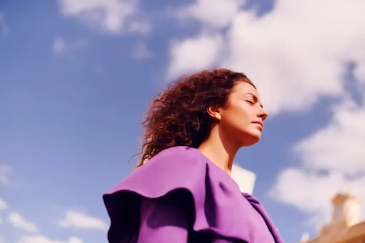 Mujer de piel blanca, pelo rizado enrojecido hacia atrás por el viento, vestida con una blusa morada. Tiene los ojos cerrados, de lado. En el fondo cielo azul y nubes.
