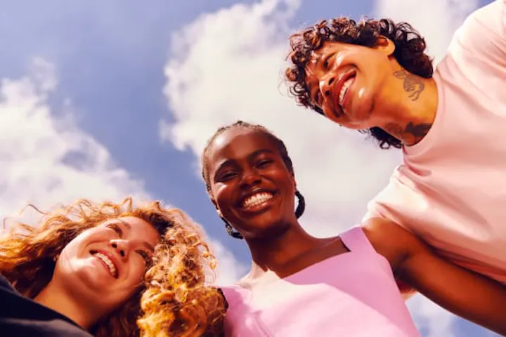 Tres personas en la imagen. A la izquierda, rostro de una mujer de piel blanca y cabello rubio rizado. En el centro, mujer de piel negra y cabello corto trenzado, vistiendo una camiseta rosa. A la derecha, el rostro de un hombre de piel rojiza con cabello rizado. Todos están sonriendo con un cielo azul con nubes en el fondo. 