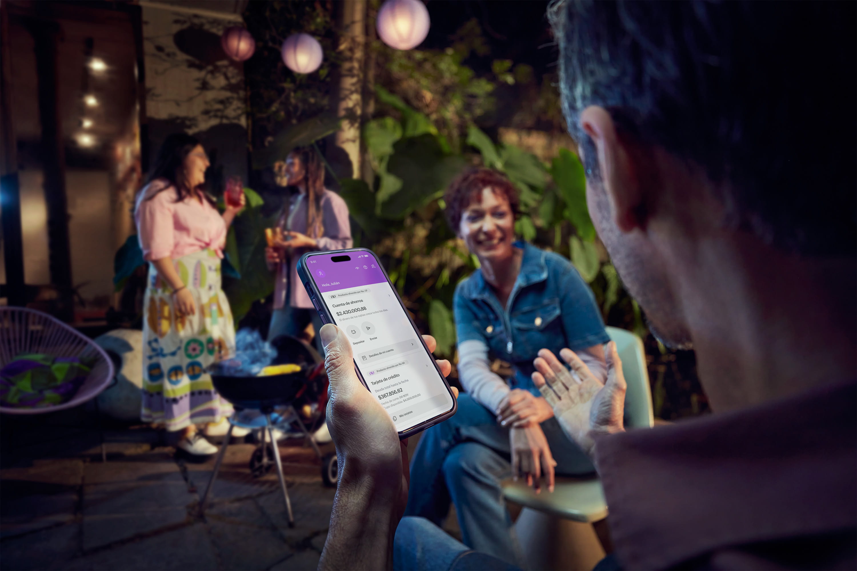 Smartphone con la app de Nu sostenido por un hombre que conversa con una mujer en una fiesta de asado en casa. Smartphone con la app de Nu sostenido por un hombre que conversa con una mujer en una fiesta de asado en casa.