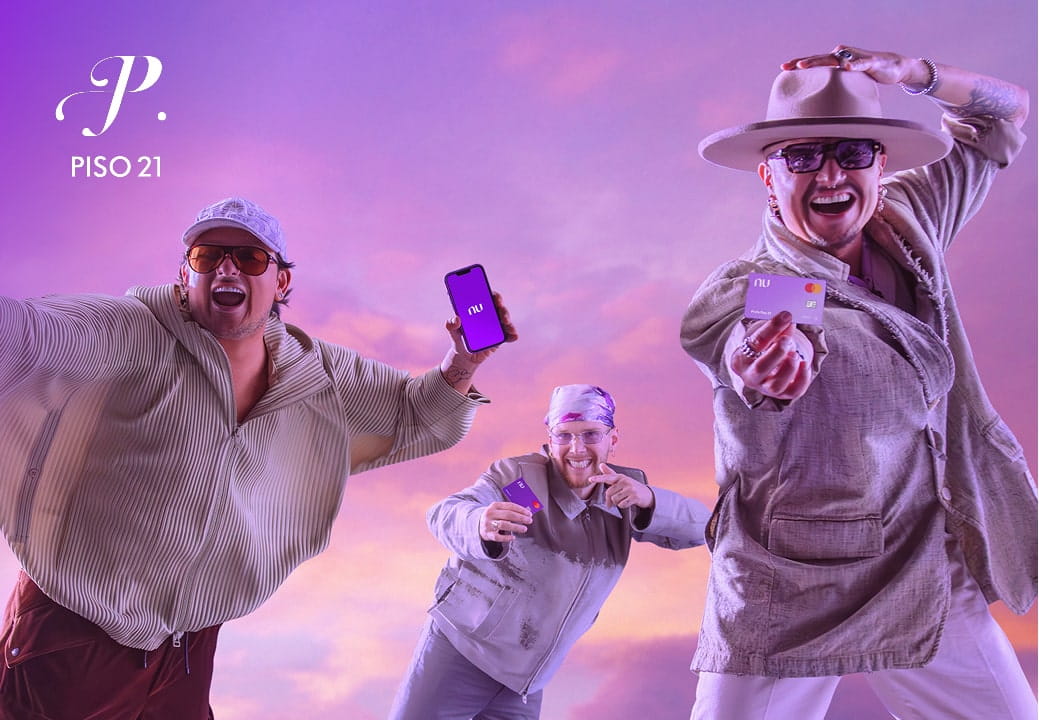 Tres integrantes del grupo musical Piso 21 posan para la cámara, con fondo de atardecer morado. Uno de ellos, en el centro, sostiene un teléfono morado con la aplicación de Nubank abierta, mientras los otros dos, en los costados, sostienen tarjetas moradas de Nubank. Tres integrantes del grupo musical Piso 21 posan para la cámara, con fondo de atardecer morado. Uno de ellos, en el centro, sostiene un teléfono morado con la aplicación de Nubank abierta, mientras los otros dos, en los costados, sostienen tarjetas moradas de Nubank.