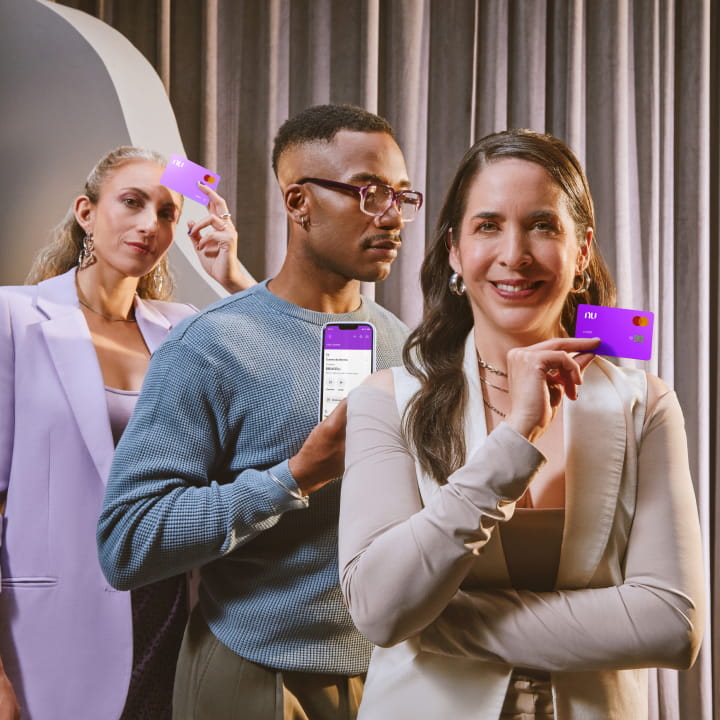 Dos mujeres y un hombre, la primer mujer sosteniendo la terjeta de débito Nu, la segunda mujer sosteniendo la tarjeta de crédito Nu y el hombre sosteniendo un teléfono con la aplicación de Nu abierta